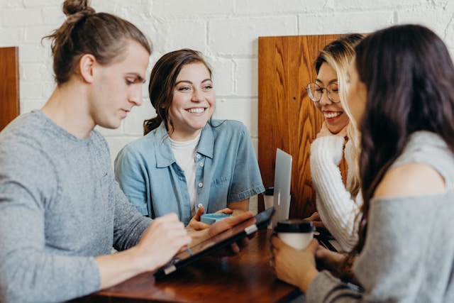 Travellers talking with a host across a table, one holding a tablet.